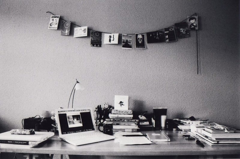 The picture shows a desk with an open laptop, wall decorations, books and papers spread and stacked across the table.
